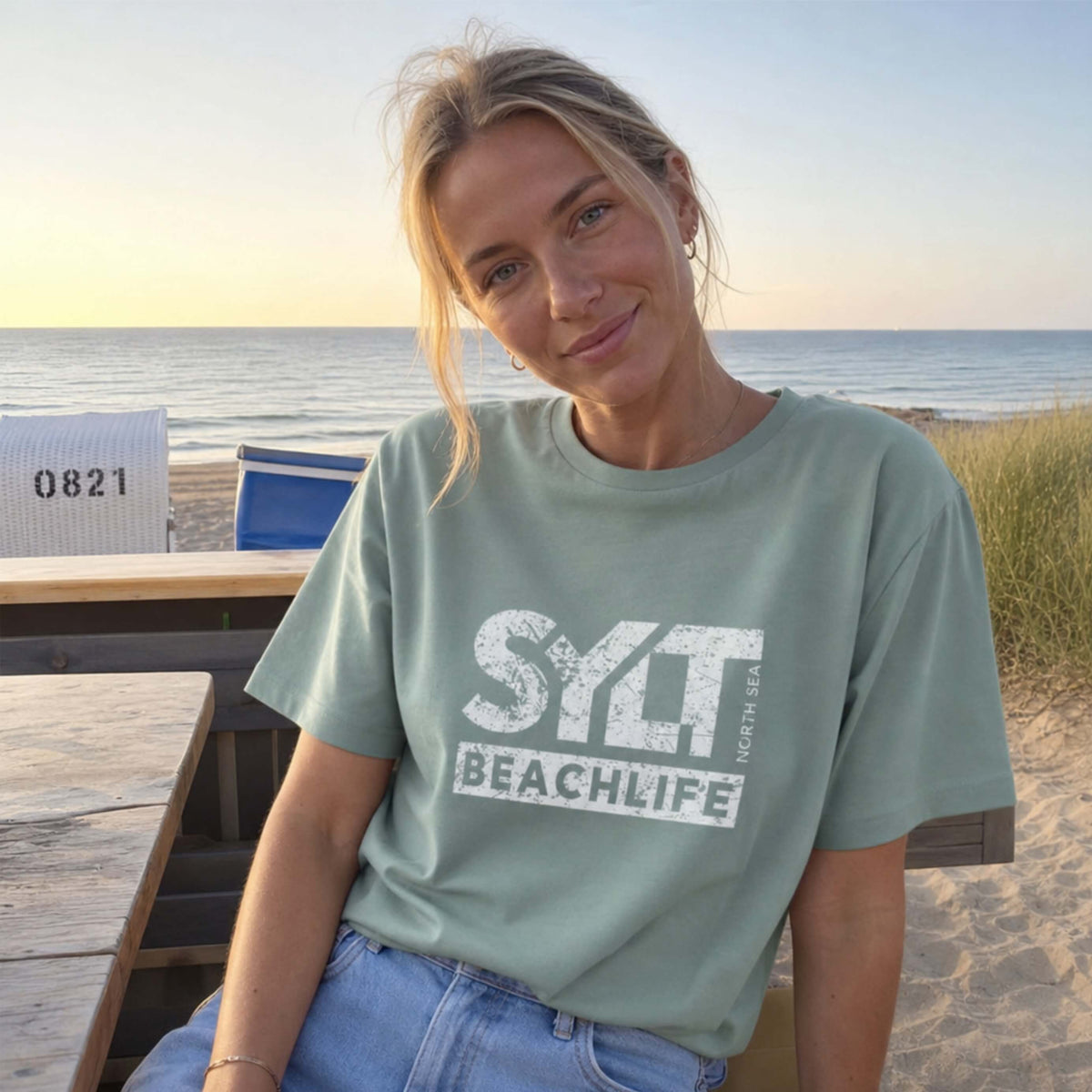 Frau in aloe farbenden Sylt T-Shirt steht auf sylt. Im Hintergrund sieht man Dünen, Strandkörbe am Strand und die Nordsee.