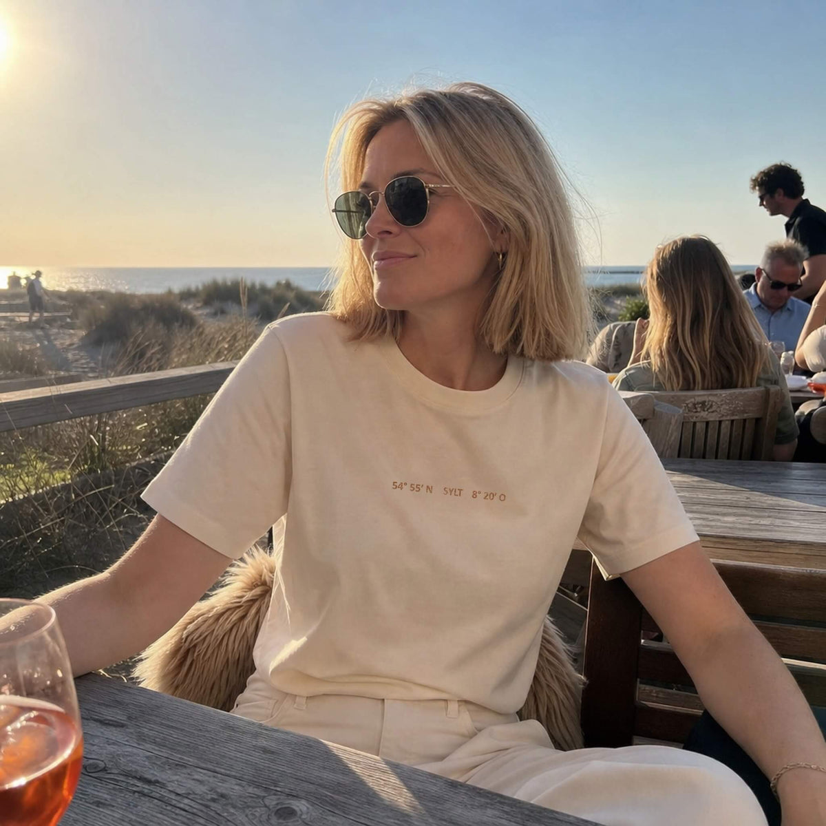 Frau sitzt lässig in einer Strand-Location und trägt dabei ein hellbeiges T-Shirt mit einem kleinen Aufdruck der Sylt Koordinaten.