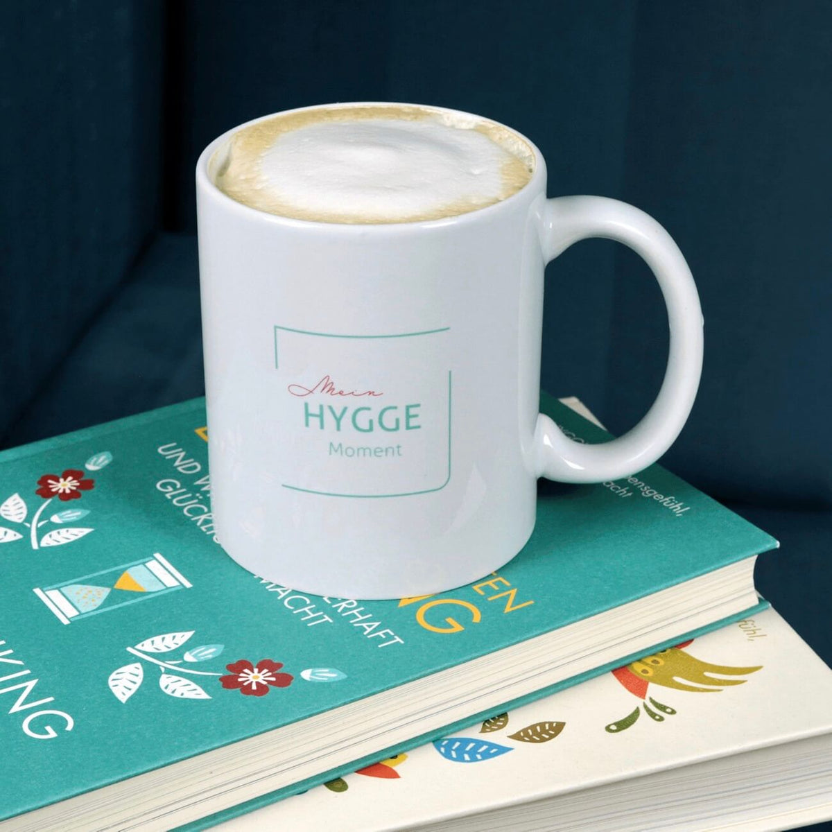 Weißer Keramik-Becher mit Aufdruck Mein Hygge Moment steht auf zwei Büchern.