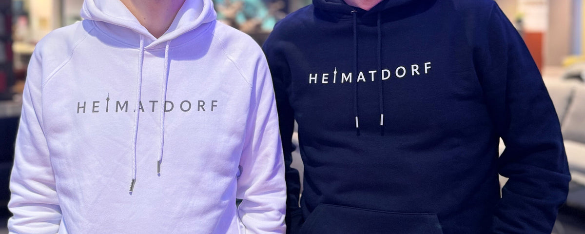 Zwei Düsseldorf Hoodies, einmal in weiß mit grauem Aufdruck Heimatdorf und einmal in dunkelblau mit weißem Heimatdorf Aufdruck.