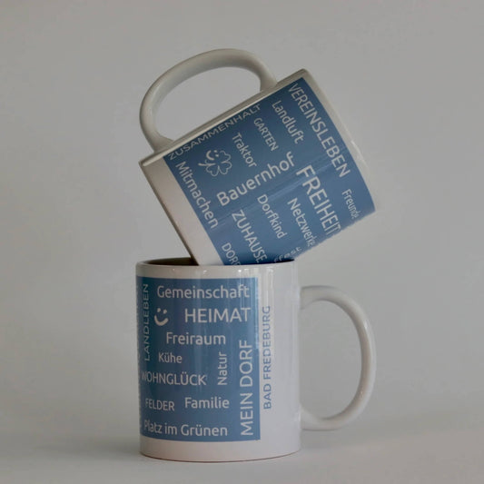 Heimatbecher aus weißer Keramik mit blauer Banderole und typischen Heimatbegriffen als Aufdruck. Mit Dorfnamen individualisierbar. 
