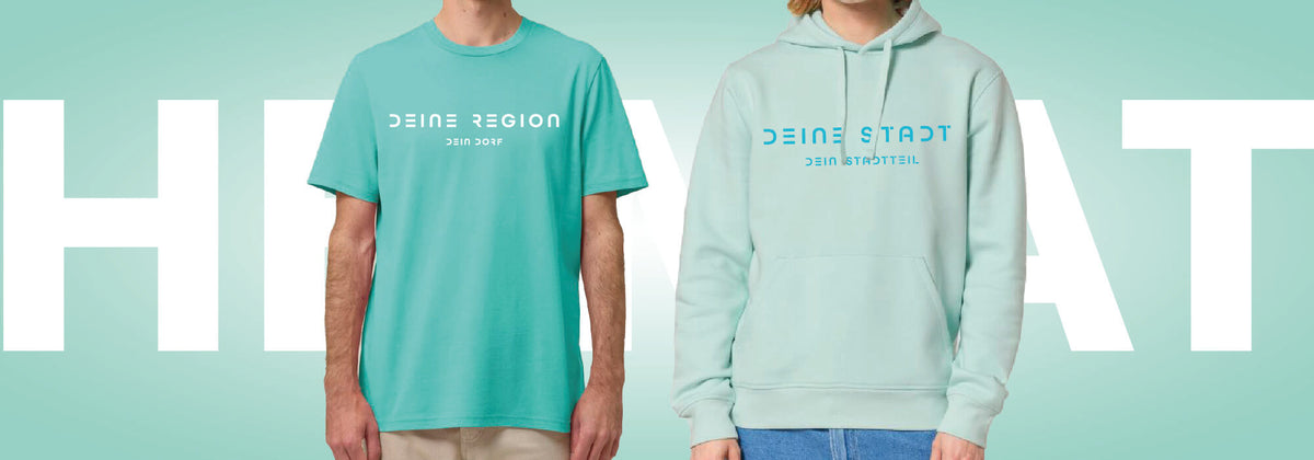 Heimat T-Shirt in türkis und Heimat-Hoodie in helltürkis werden vor einem weißen Heimat-Schriftzug präsentiert. Als Aufdruck kann entweder die Region und ein Dorf oder der Name einer Stadt und eines Stadtteils individuell gewählt werden. Die Schrift des Aufdrucks ist sehr modern mit Cut-Outs.