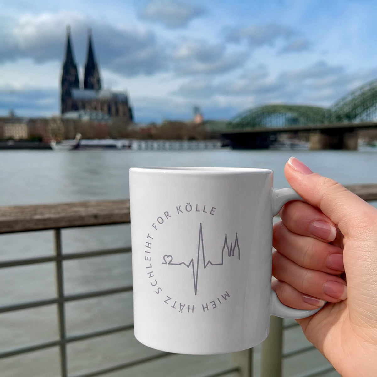 Weißer Becher mit Mieh Hätz schleiht for Kölle Motiv wird von einer Frauen-Hand gehalten. Im Hintergrund sieht man den Rhein und den Kölner Dom.