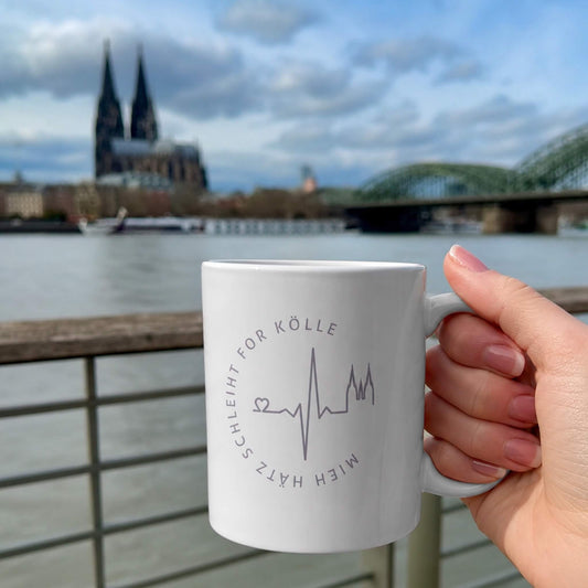 Weißer Becher mit Mieh Hätz schleiht for Kölle Motiv wird von einer Frauen-Hand gehalten. Im Hintergrund sieht man den Rhein und den Kölner Dom.