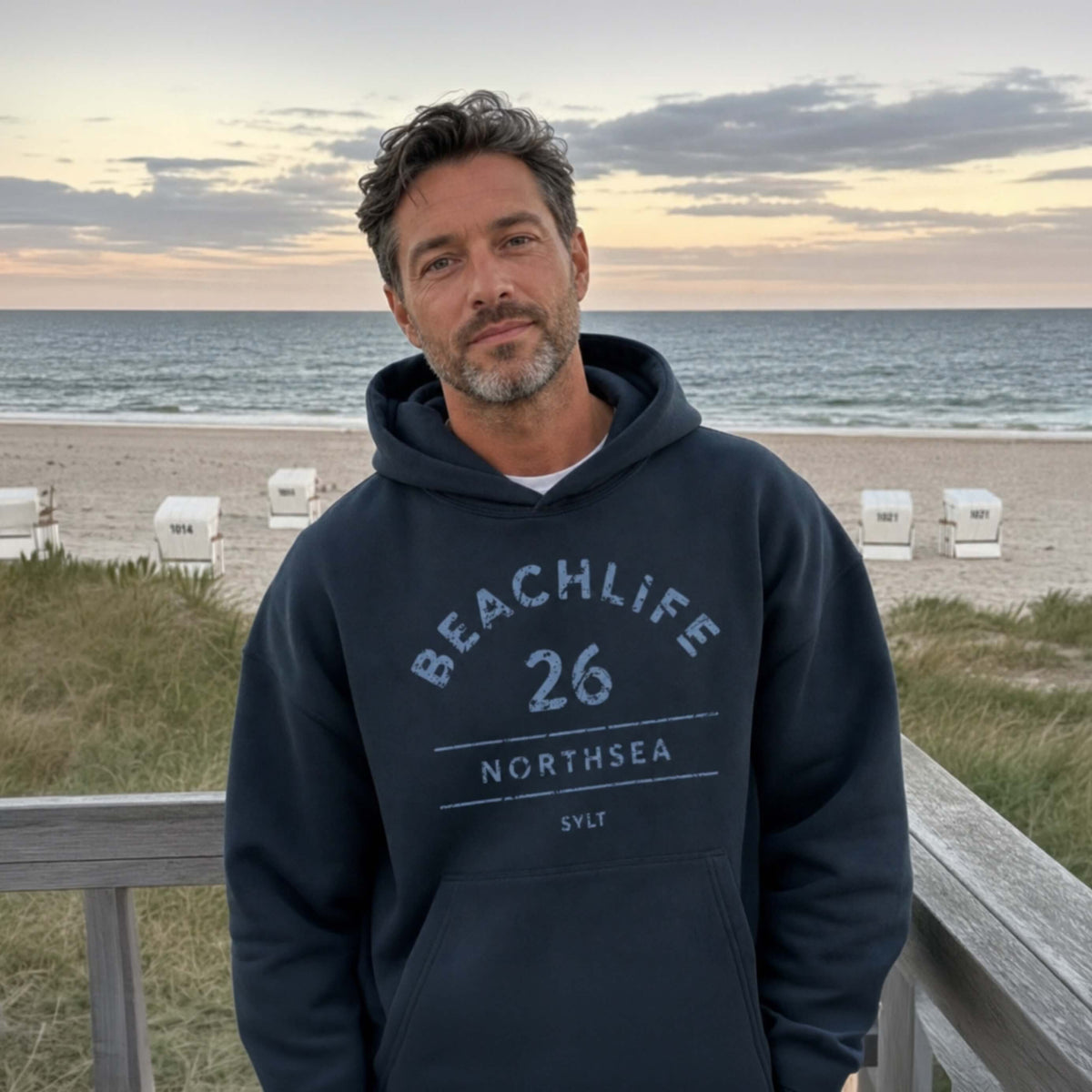 Braunhaariger Mann  in graublauen Hoodie mit "BEACHLIFE 26 NORTHSEA SYLT" Aufdruck in entspannter Strandszenerie.
