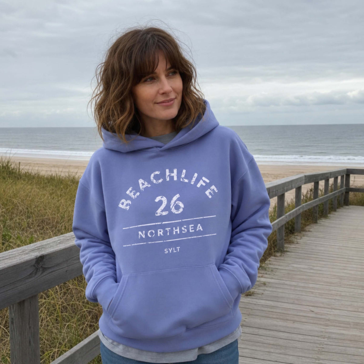 Braunhaarige Frau in violetem Hoodie mit "BEACHLIFE 26 NORTHSEA SYLT" Aufdruck in entspannter Strandszenerie.
