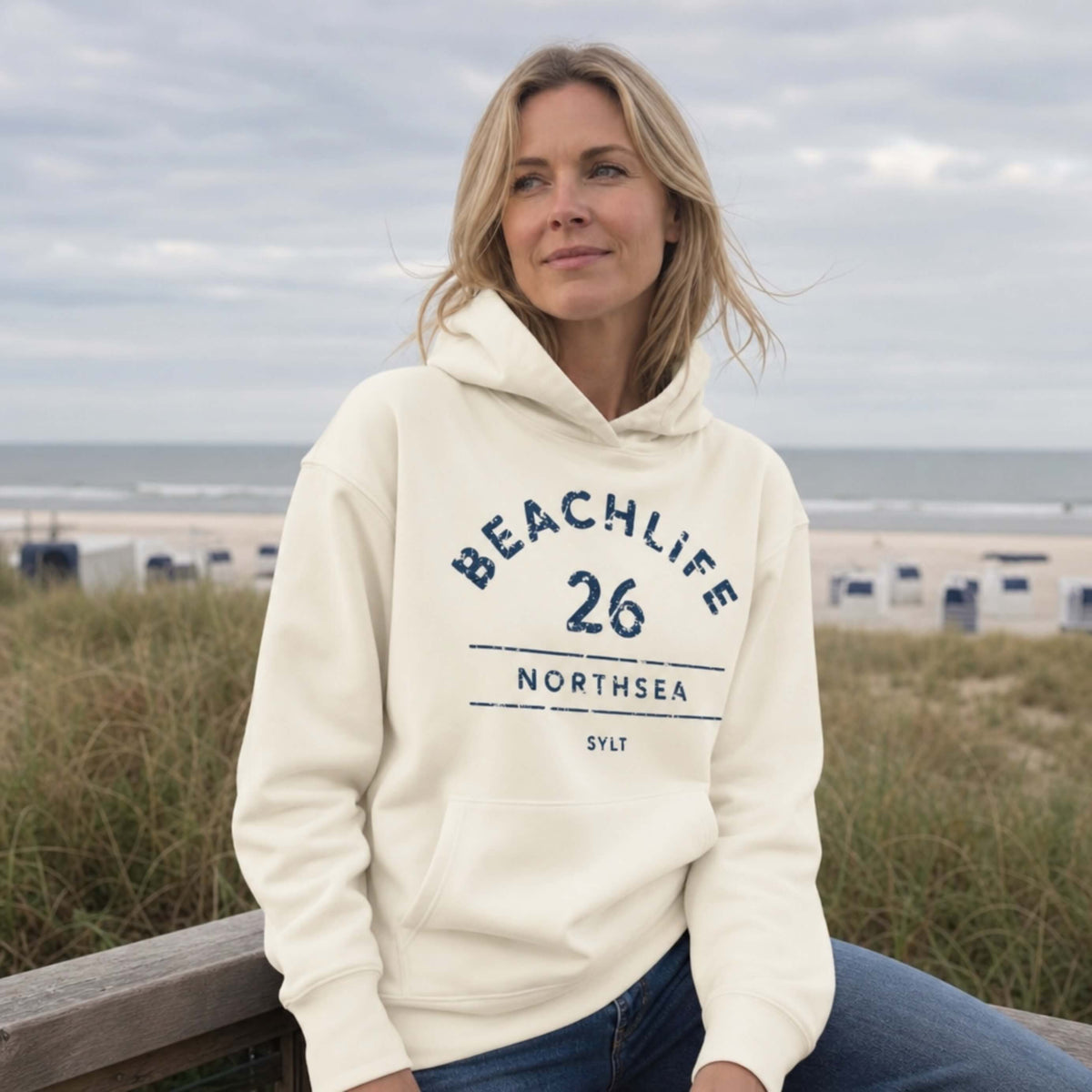 blonde Frau in vanille farbenden Hoodie mit "BEACHLIFE 26 NORTHSEA SYLT" Aufdruck in entspannter Strandszenerie.