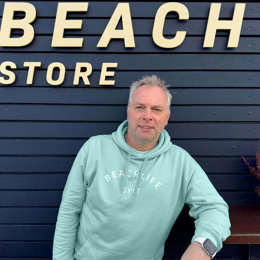 Mann steht mit einem aqua farbigem Sylt Hoodie vor einem schwarzen Strandhaus. Vorne auf dem Hoodie ist der Begriff Beachlife leicht gerundet aufgedruckt. Darunter steht mittig in weiß Sylt.