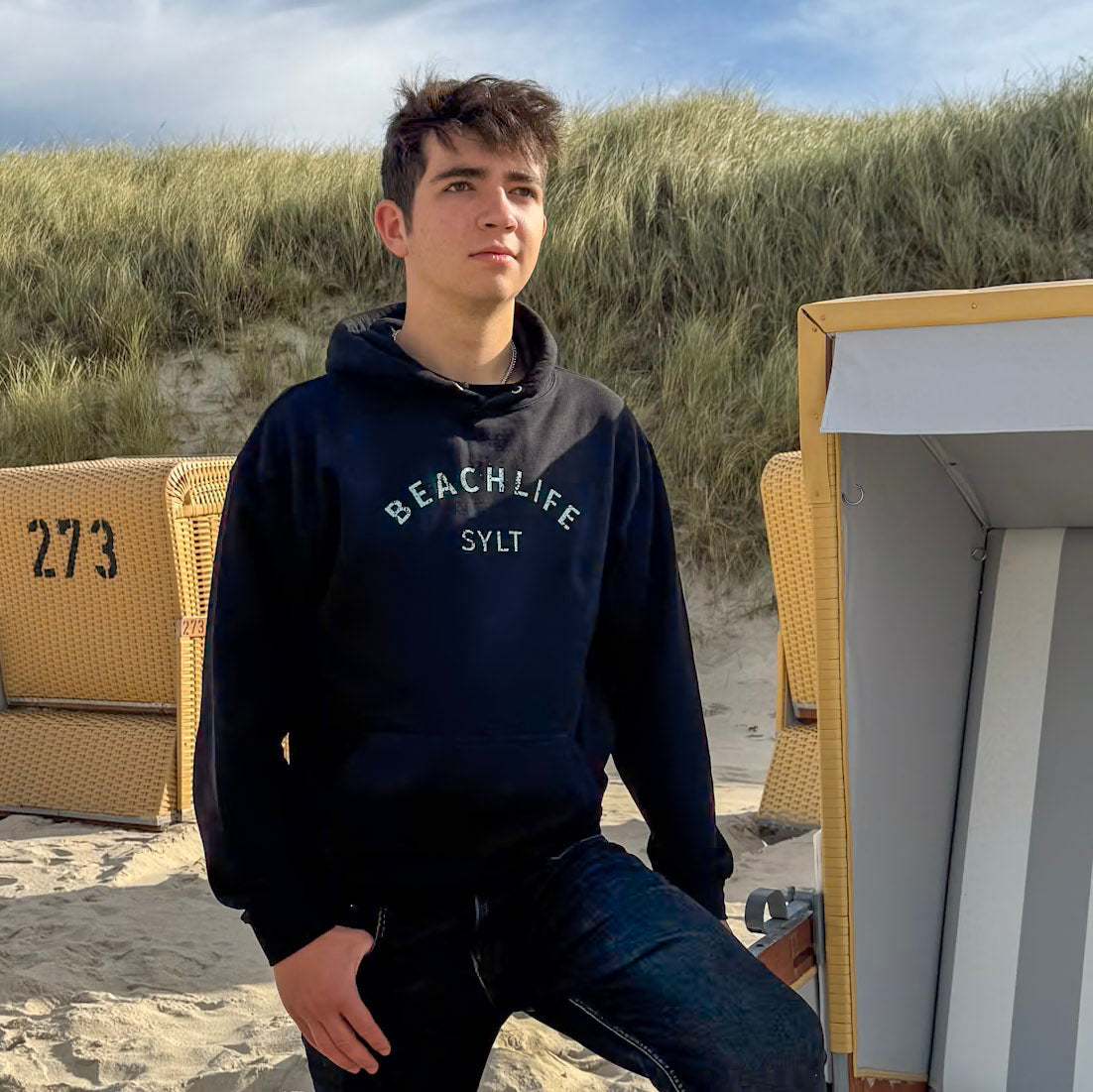 Junger Mann steht mit schwarzem Sylt Hoodie an einem Strandkorb. Mittig findet sich ein türkiser Beachlife Sylt Aufdruck.