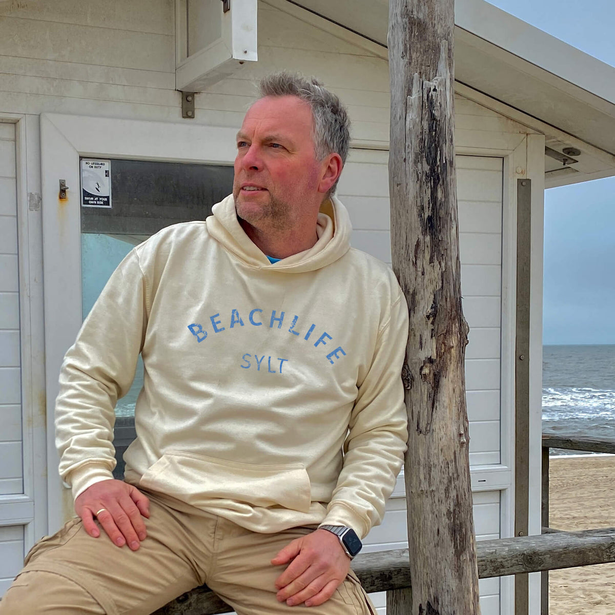 Mann sitzt lässig an einem Strandhäuschen mit Sylt Hoodie in hellbeige und blauem Aufdruck Beachlife Sylt