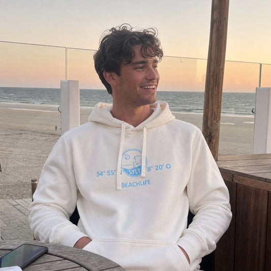 Mann steht mit Hellbeige Sylt-Hoodie vor einem Türkisen Surfbrett. Der Hoodie ist vorne in türkis mit den Koordinaten von Sylt sowie einem Strandkorb bedruckt. Darunter ist der Begriff Beschlief aufgedruckt.