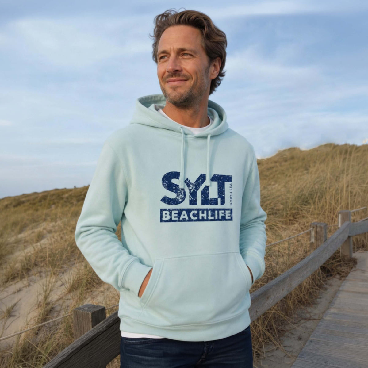 Mann in blauem Hoodie mit dunkelblauen 'SYLT NORTH SEA BEACHLIFE' Logo, steht auf Sylt.