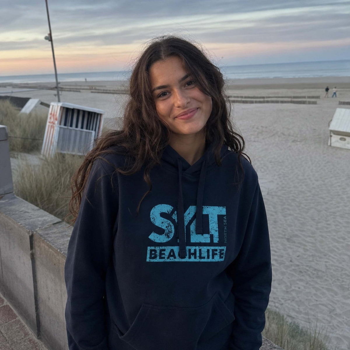 Frau in dunkelblauen Hoodie mit hellblauen 'SYLT NORTH SEA BEACHLIFE' Logo, steht auf Sylt.