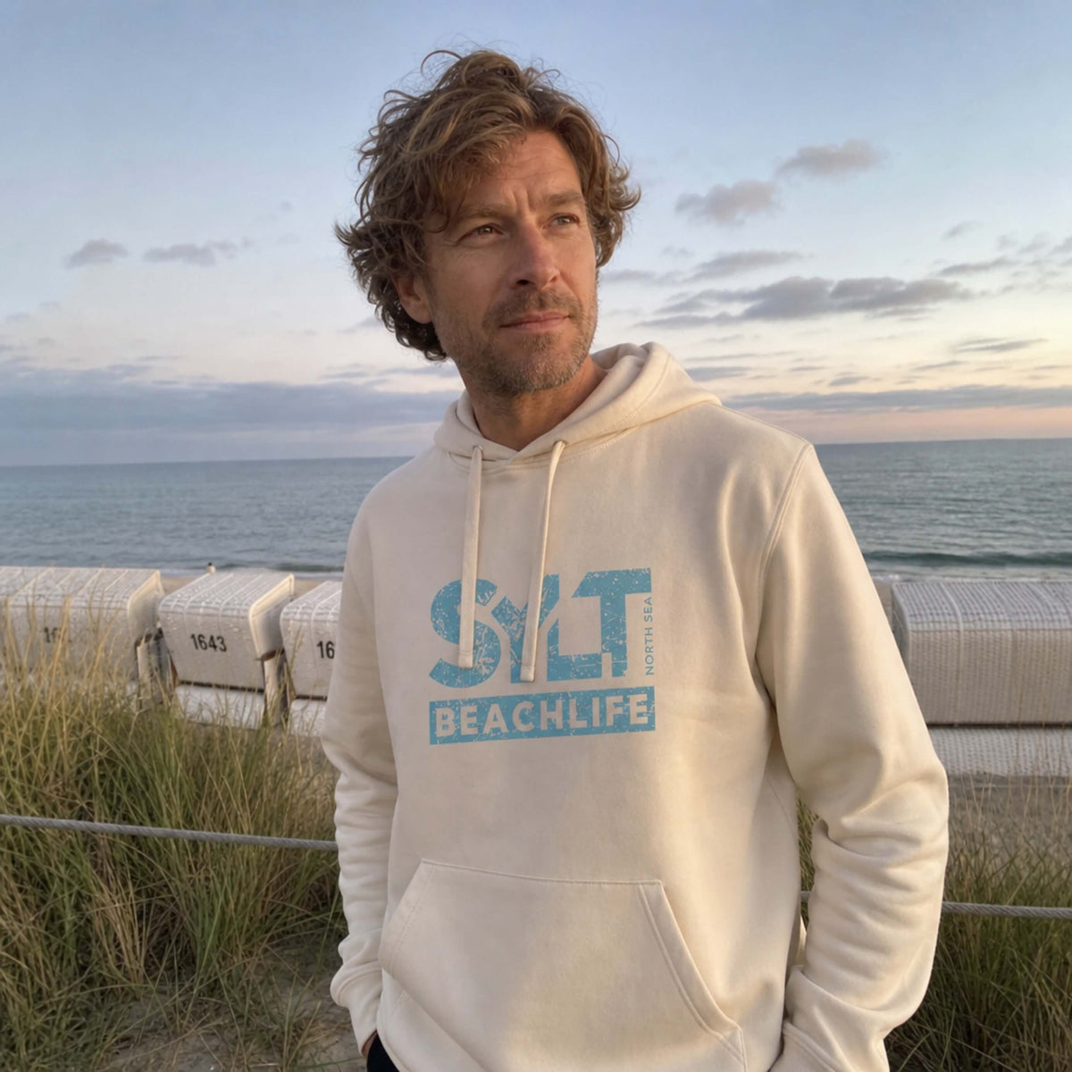 Mann in beigem Hoodie mit hellblauen 'SYLT NORTH SEA BEACHLIFE' Logo, steht auf Sylt.