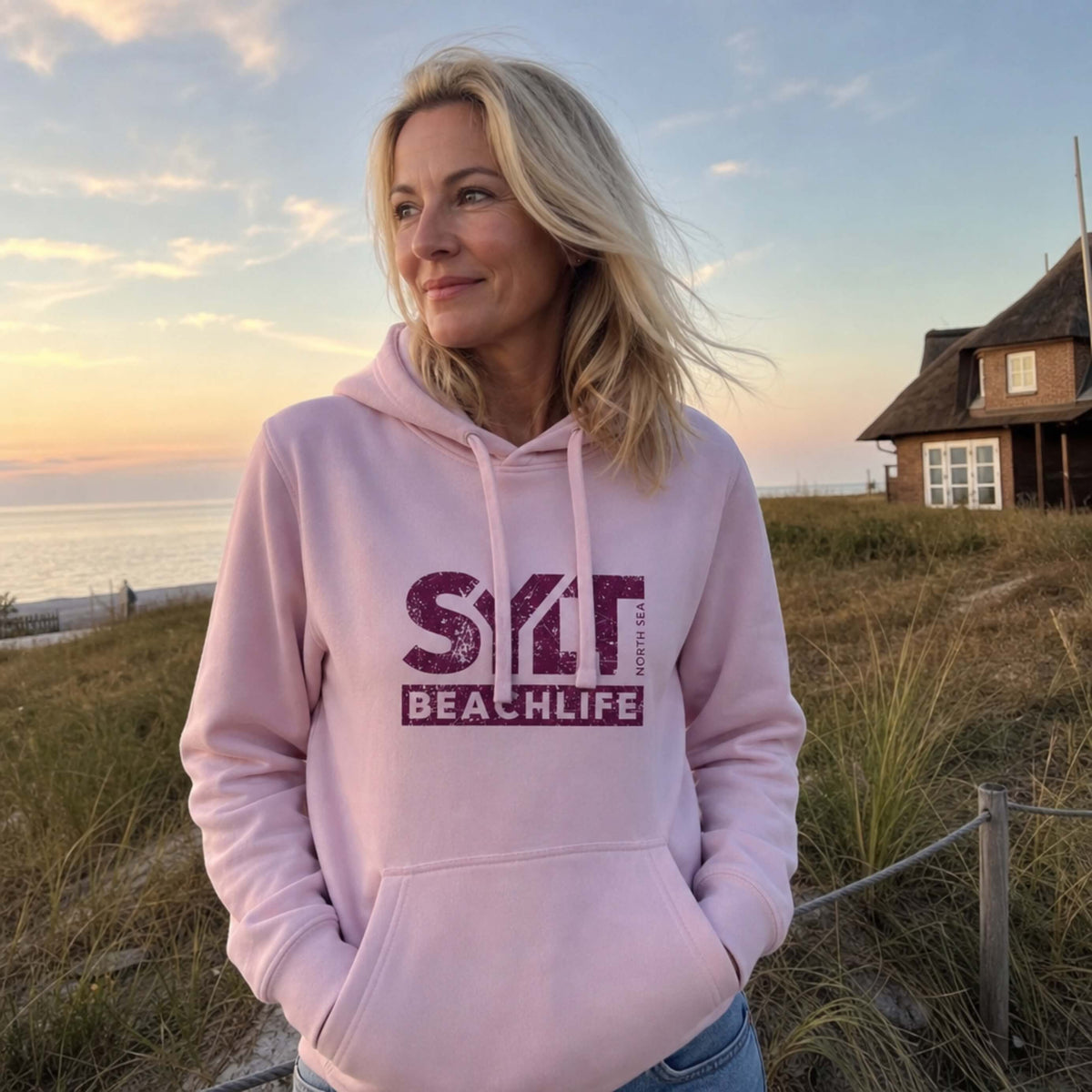Frau in rosanem Hoodie mit rosanem 'SYLT NORTH SEA BEACHLIFE' Logo, steht auf Sylt.