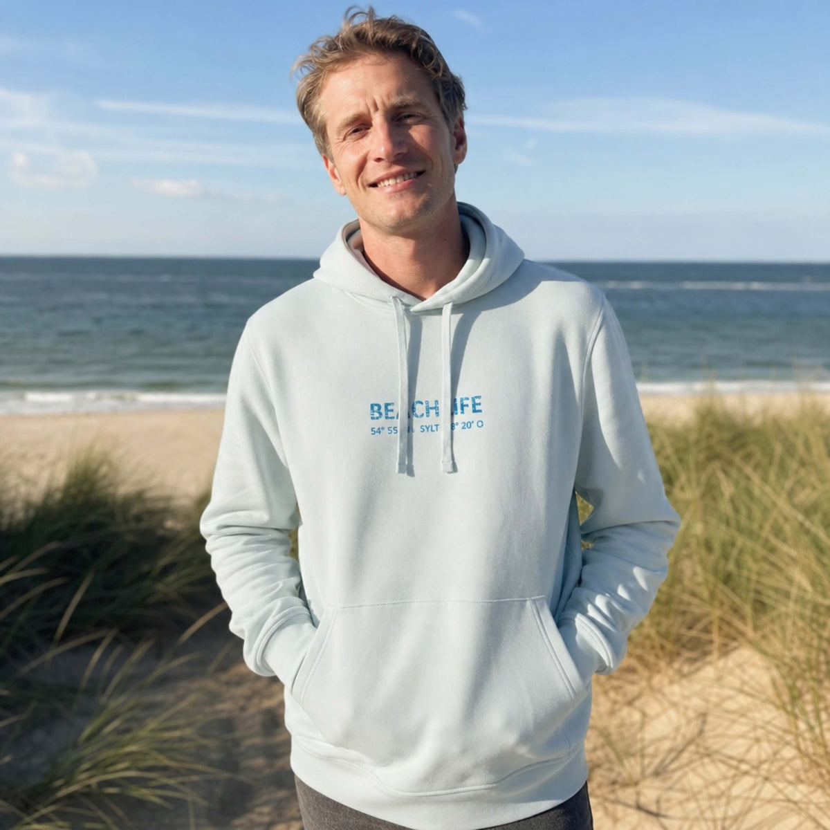 Frau steht auf Sylt in einem leichtblauen Hoodie mit blauem, mittigen Aufdruck Beachlife Sylt und den Koordinaten.