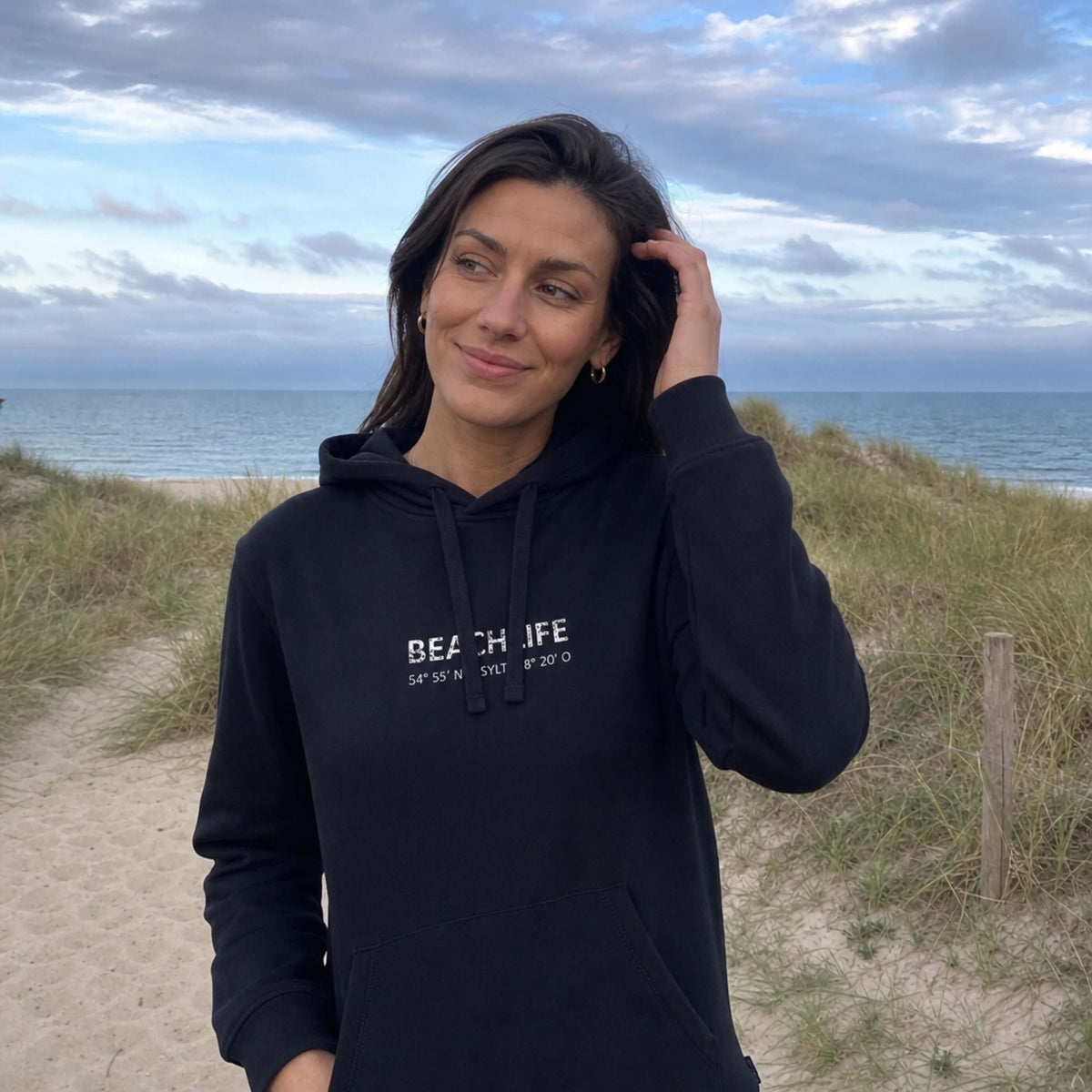 Frau steht auf Sylt in einem schwarzen Hoodie mit weißem, mittigen Aufdruck Beachlife Sylt und den Koordinaten.