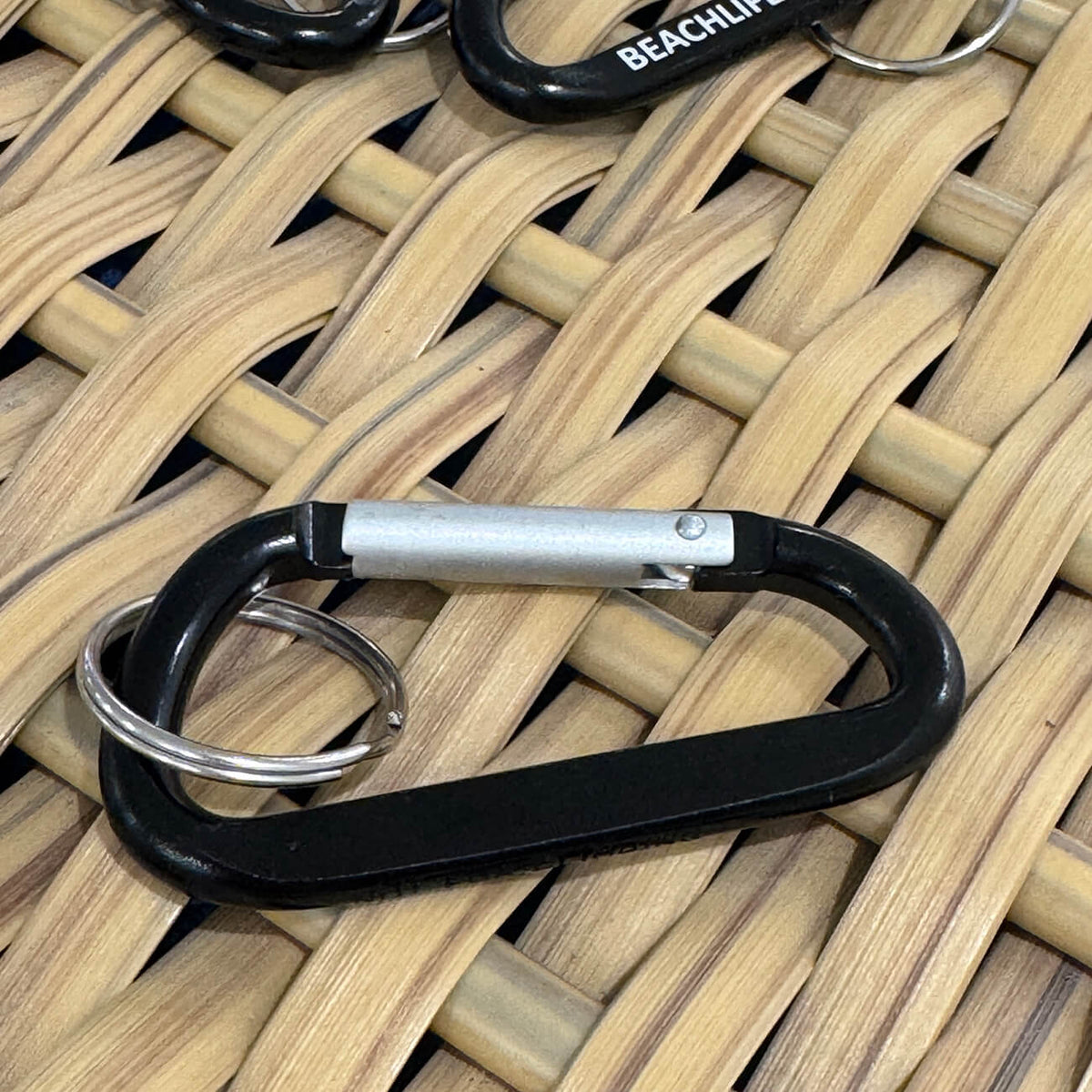 Rückseite eines schwarzen Schlüsselanhängers in Karabiner-Form, der auf der Vorderseite die Fräsung Beachlife Sylt aufweist.