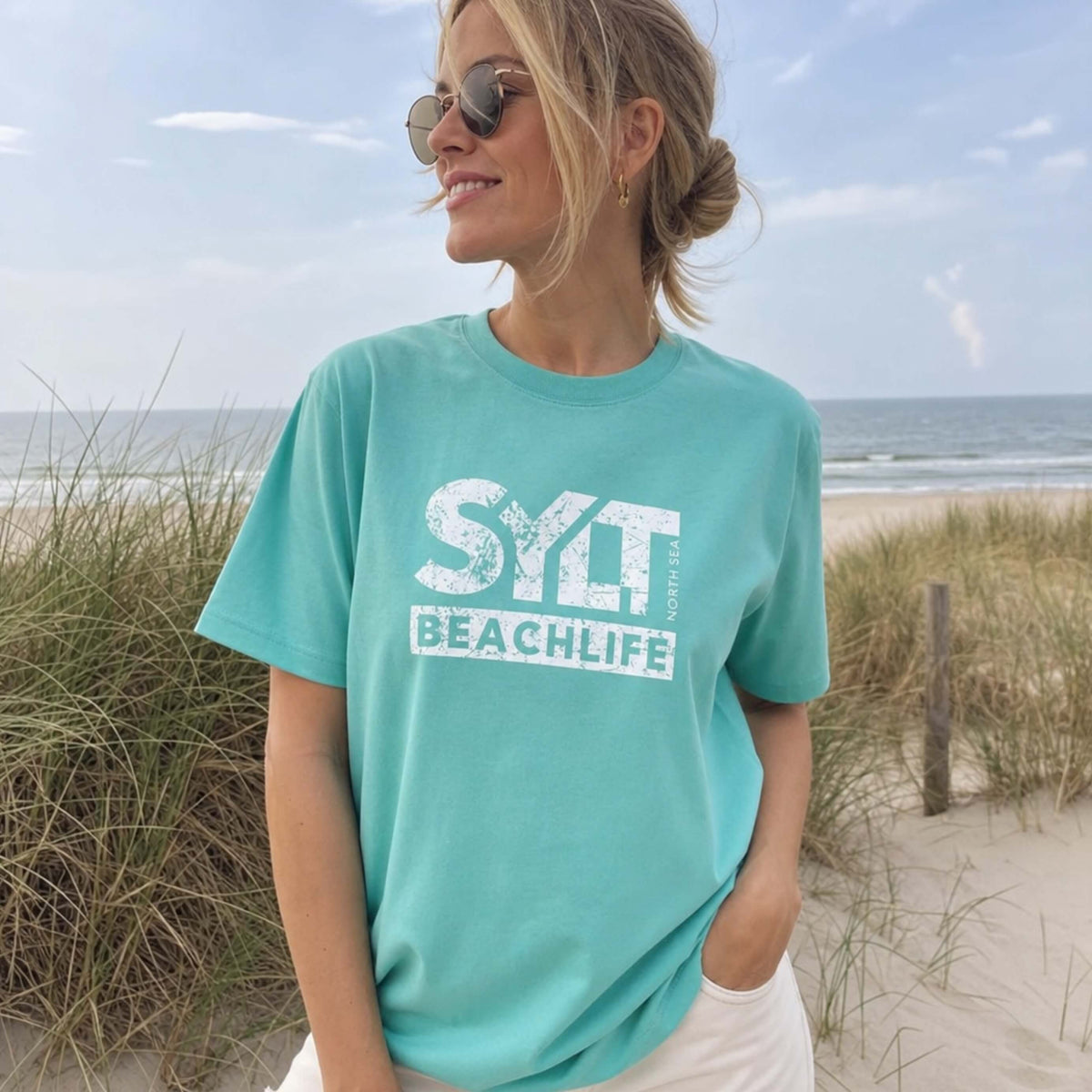 Frau in aqua farbenden Sylt T-Shirt steht auf sylt. Im Hintergrund sieht man Dünen, Strandkörbe am Strand und die Nordsee.