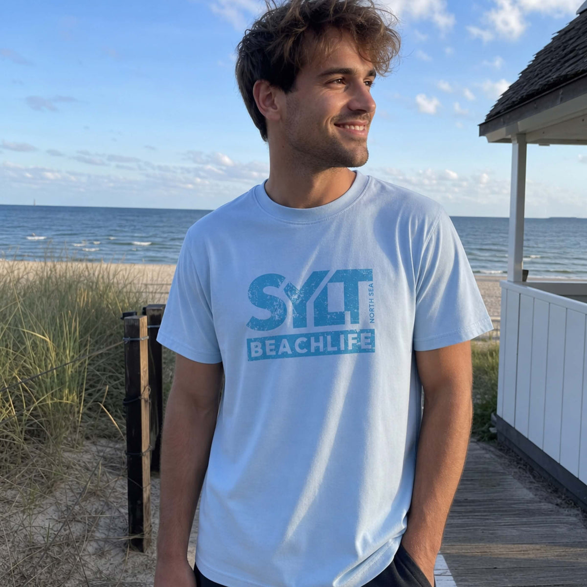 Mann in hellblau farbenden Sylt T-Shirt steht auf sylt. Im Hintergrund sieht man Dünen, Strandkörbe am Strand und die Nordsee.
