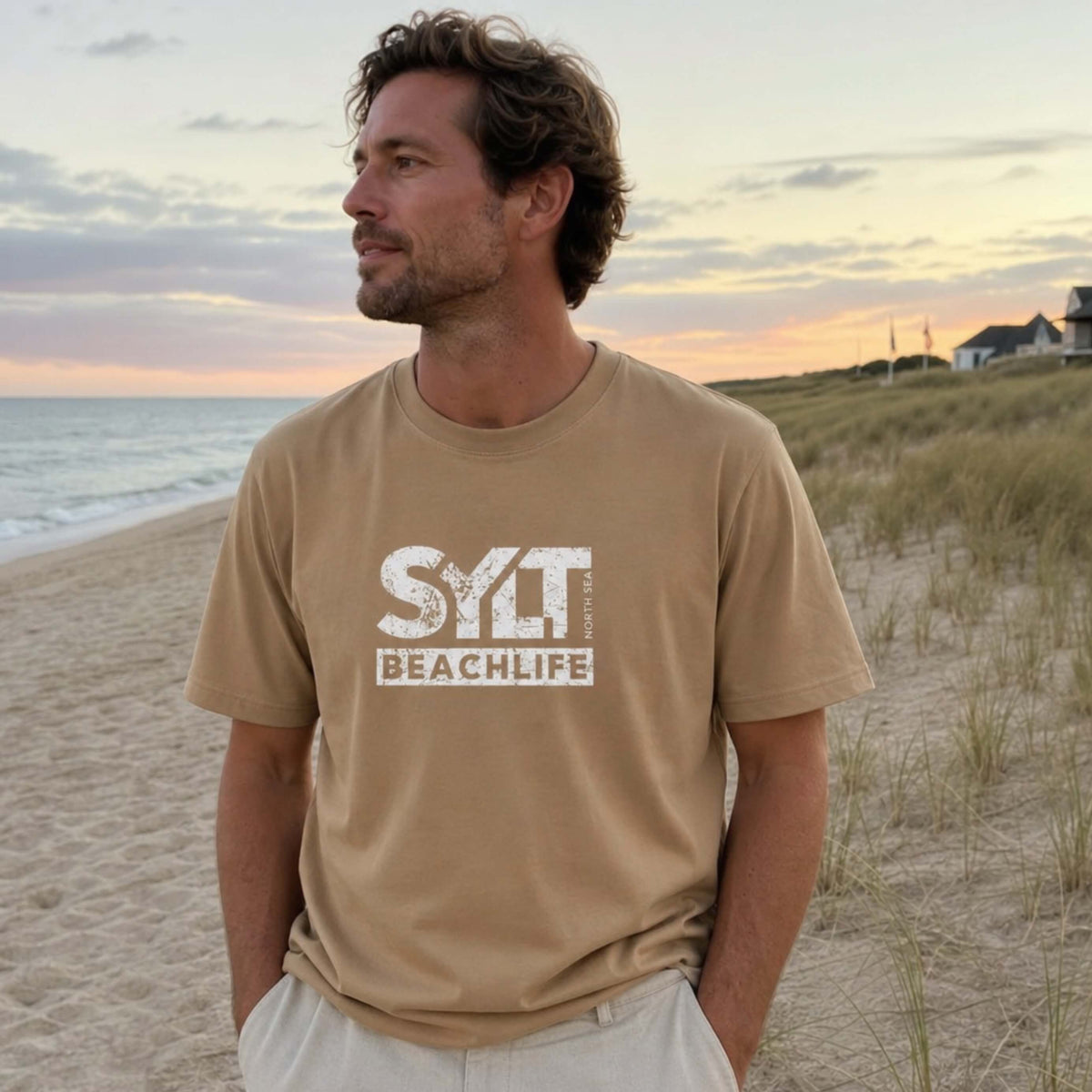 Mann in Reed farbenden Sylt T-Shirt steht auf sylt. Im Hintergrund sieht man Dünen, Strandkörbe am Strand und die Nordsee.