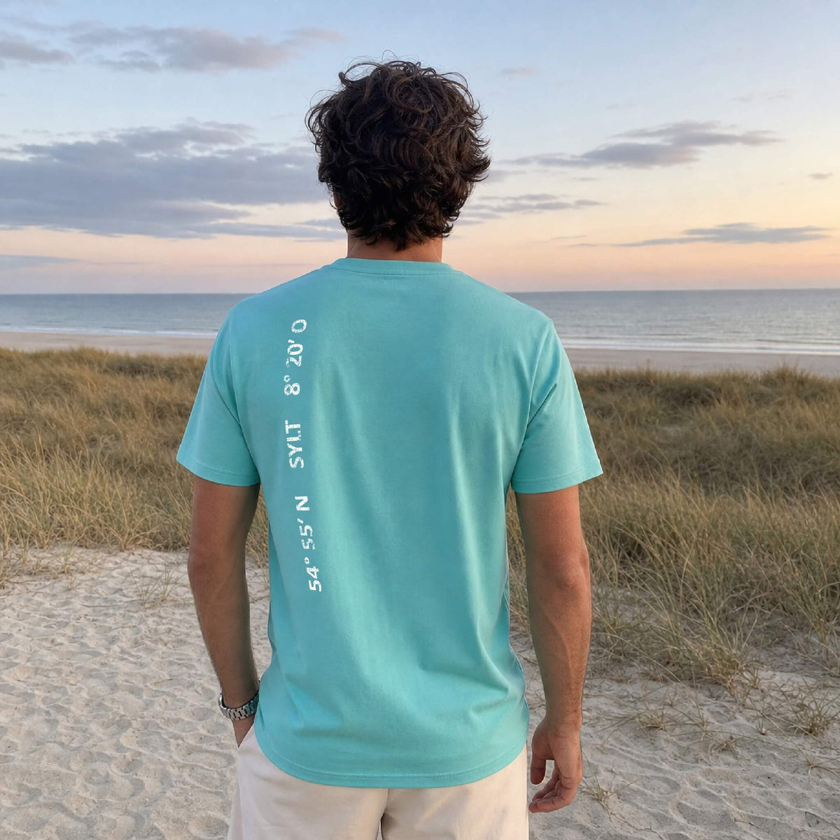 Mann steht mit türkisem T-Shirt am Strand und guckt aufs Meer. Auf seinem Rücken befindet sich ein weißer Aufdruck mit den Sylter Koordinaten.