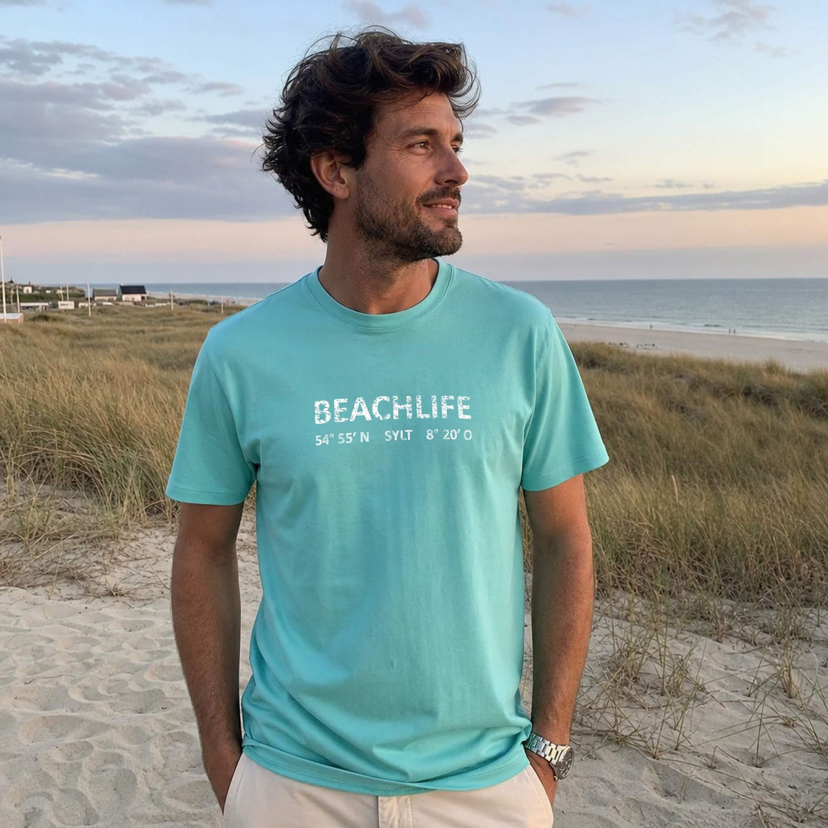 Mann steht mit türkisem Sylt T-Shirt und weißem Aufdruck Beachlife Sylt am Strand.