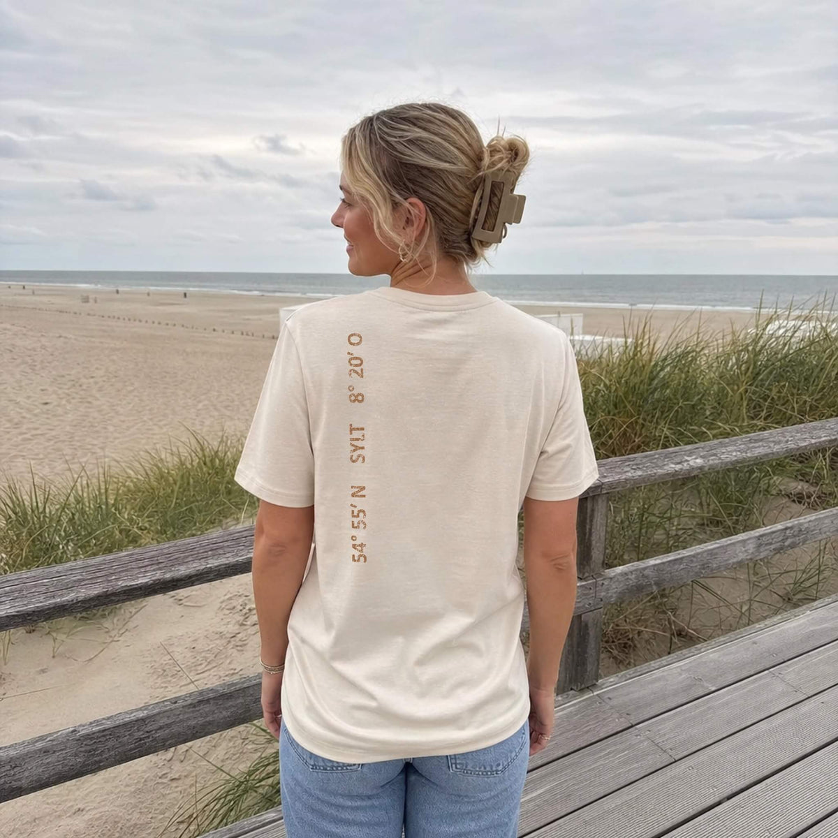 Frau steht mit einem beigen Sylt-T-Shirt in den Dünen und schaut aufs Meer. Auf der linken Schulter sieht man senkrecht einen kupferfarbigen Aufdruck der Sylt Koordinaten