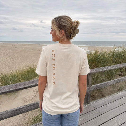Frau steht mit einem beigen Sylt-T-Shirt in den Dünen und schaut aufs Meer. Auf der linken Schulter sieht man senkrecht einen kupferfarbigen Aufdruck der Sylt Koordinaten