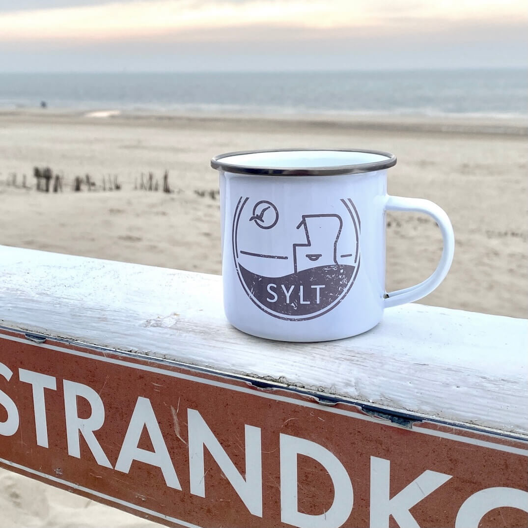 Weißer Emaillebecher mit Sylt-Aufdruck steht auf einer weißen Brüstung in Westerland. Im Hintergrund sieht man den Stand und das Meer. Der weiße Emaillebecher ist in dunkelbeige bedruckt. Das Motiv ist ein Sylter Nordseestrandkorb, eine Möwe die durch die Sonne fliegt sowie der Begriff Sylt.