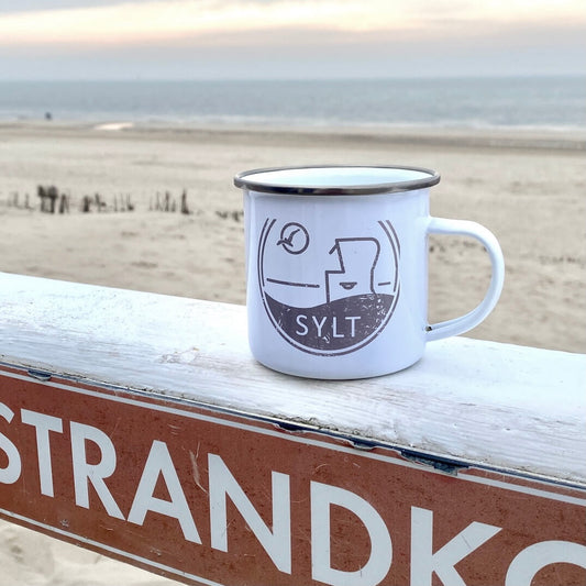 Weißer Emaillebecher mit Sylt-Aufdruck steht auf einer weißen Brüstung in Westerland. Im Hintergrund sieht man den Stand und das Meer. Der weiße Emaillebecher ist in dunkelbeige bedruckt. Das Motiv ist ein Sylter Nordseestrandkorb, eine Möwe die durch die Sonne fliegt sowie der Begriff Sylt.