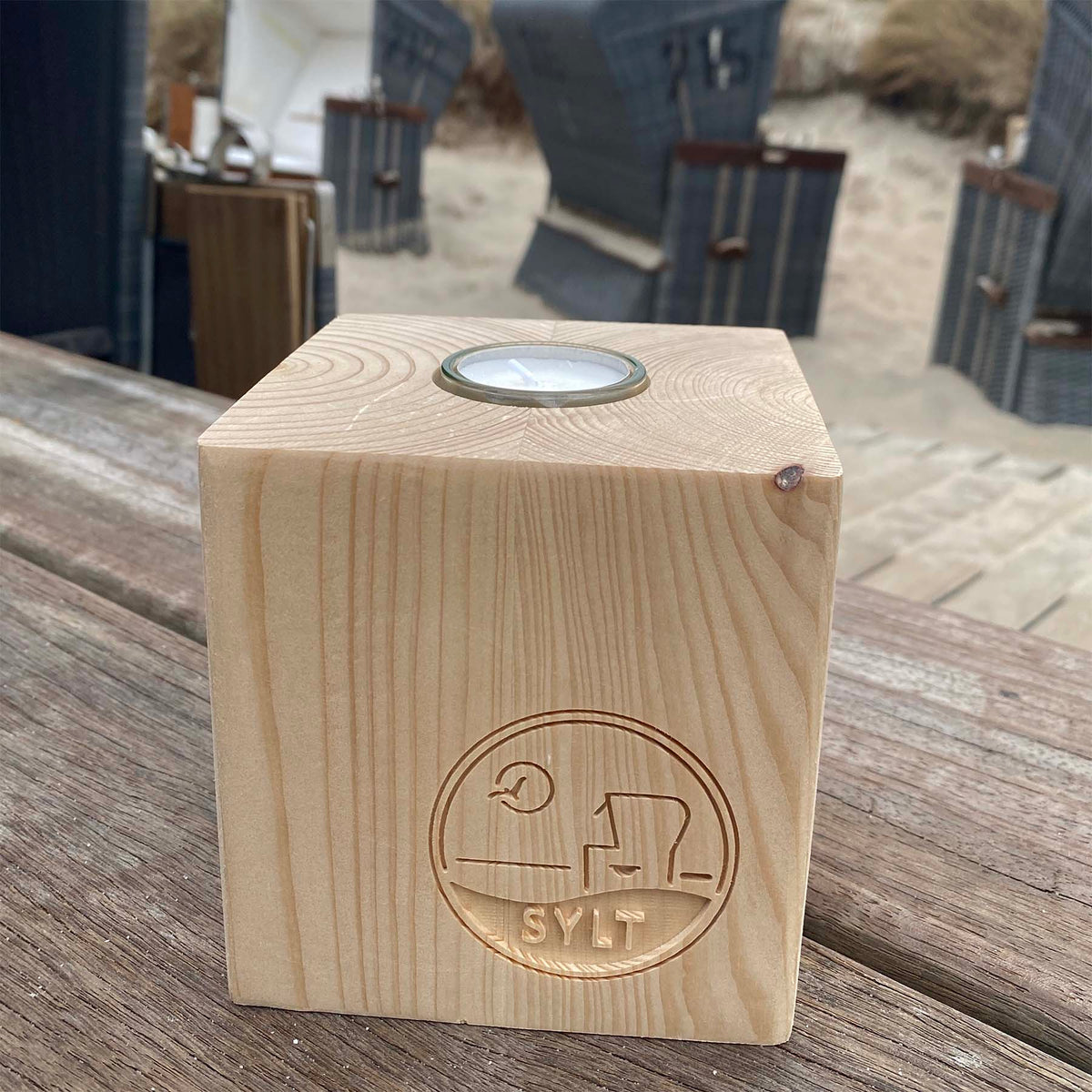 Holz Teelichtwürfel steht in Strandbistro auf einem rustikalen Holztisch. Im Hintergrund sind Strandkörbe zu sehen. Der Teelichtwürfel hat eine Fräsung mit dem Begriff Sylt, einem Strandkorb sowie einer Möwe, die durch die Sonne fliegt. 