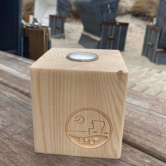 Holz Teelichtwürfel steht in Strandbistro auf einem rustikalen Holztisch. Im Hintergrund sind Strandkörbe zu sehen. Der Teelichtwürfel hat eine Fräsung mit dem Begriff Sylt, einem Strandkorb sowie einer Möwe, die durch die Sonne fliegt. 