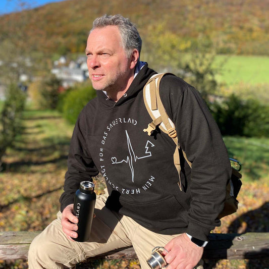 Mann sitzt mit schwarzem Hoodie und Rucksack im Sauerland auf einem Holzbalken. Auf dem Hoodie ist in grau und zeigt einen Herzschlag der mit der Grafik Herz anfängt und mit einer grafik von  einem Tannenbaum aufhört. Drumherum ist in rund geschrieben: Mein Herz schlägt für das Sauerland.