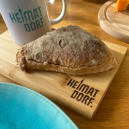 Auf einem hochwertigen Holzbrettchen liegt ein Brötchen und steht eine Tasse mit dem HeimatDorf Logo Düsseldorf. Das Frühstücksbrettchen hat unten rechts eine Lasergravur mit dem Heimatdorf Logo. Das i ist durch den Rheinturm dargestellt.