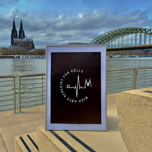 Ein Bilderrahmen mit einem schwarzen Poster steht am Rhein in Köln auf einer Stufe aus Sandstein. Im Hintergrund sieht man den Kölner Dom und die Deuter Brücke. Das Poster ist schwarz mit weißem Aufdruck. In rund steht der Schriftzug Mieh Hätz schleiht for Kölle. In der Mitte ist ein Herzschlag zu sehen, der mit einem Herz beginnt und mit einer Grafik vom Kölner Dom endet.